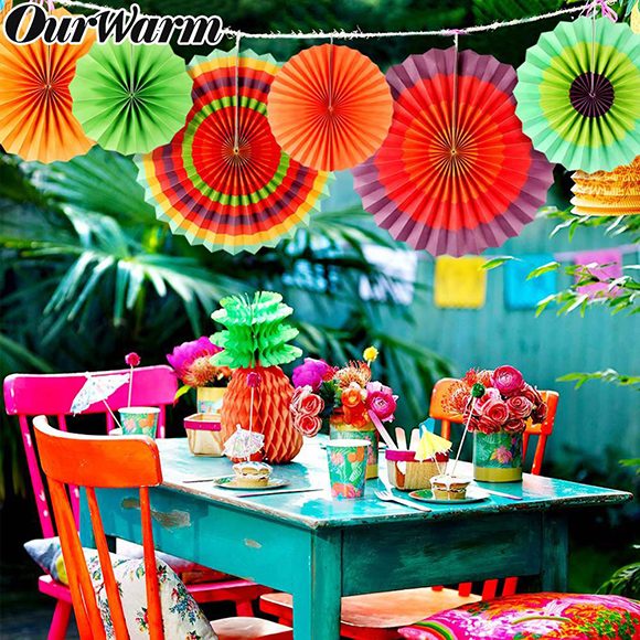 Photo of a blue-green table and pink and red painted chairs in a backyard. The table is dressed with a paper pineapple decoration and paper cups and plates, plus a can with fresh flowers. Hanging above the table is a strand of large and small unfolded circular paper decorations, which could be used for a Mexican fiesta. Green plants and a blue-green fence are slightly out of focus in the background.