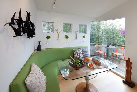 Another view of Kimball's tiny house interior, showing the lime green custom curved banquette, a custom adjustable-height glass dining table, and outside on the patio, two orange chairs and a pedestal table.