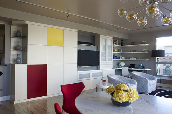 After the renovation, now the space features a wall of storage with a square in yellow and a rectangle in red, while all the other cabinet surfaces are white. Open shelving displays 3 vases at the end of the storage wall on the left. A wall-mounted television is included to the right, and to the right of that, frosted glass covers additional storage doors and drawers. Open shelving and a corner desk are at the far right on the rear wall. Next to it on the right is an open, bright window. In front of that are two grey patterned tub chairs. Back in the center front of the image is a marble patterned dining table dressed with yellow flowers, and red modern dining chairs. Above are some glass bubbles with yellow bulbs inside, hanging as a chandelier above the dining table.