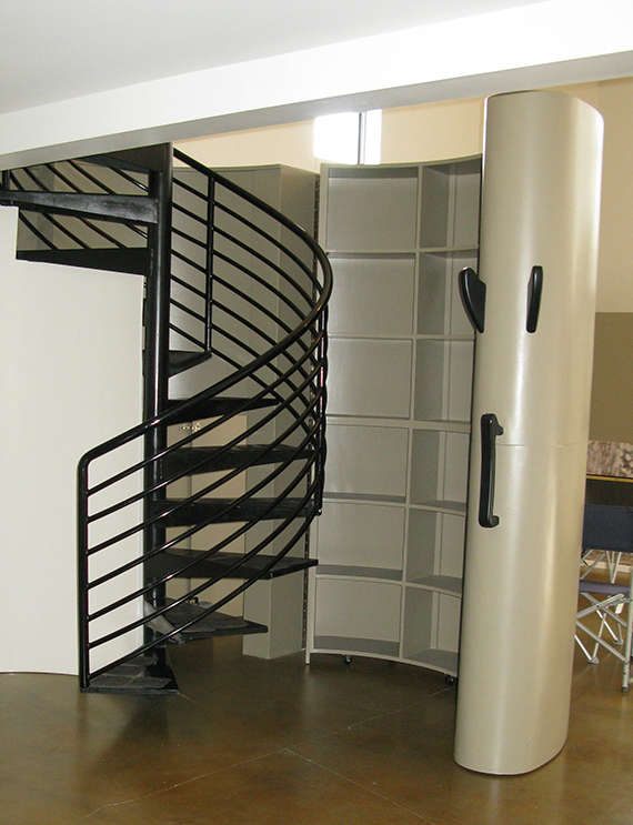 Before photo of a black metal circular staircase with a white rolling bookcase that closes around the stairs, with black handles to pull the bookcase open and shut. The outside of the rolling case resembles a white column. The flooring is a tan polished concrete, with stark white walls beyond.
