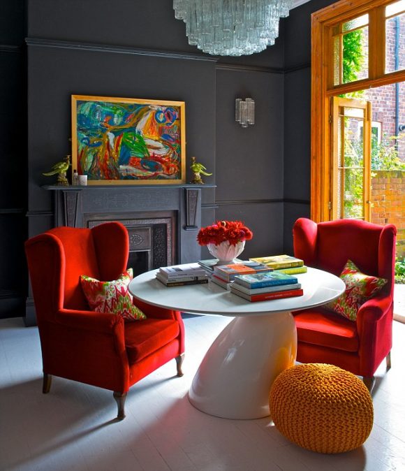 A split complementary colored living room with red wingback chairs, a white table dressed with colorful books, an orange pouf as a footrest, orange window frame, a dark grey back wall with a fireplace and a colorful framed piece of art sitting on the mantle, with a glass chandelier hanging overhead.