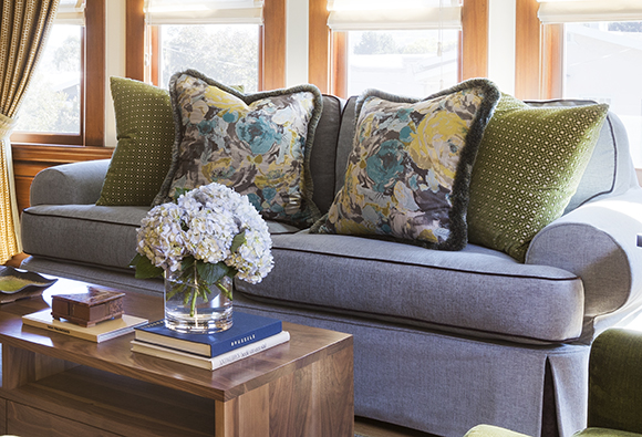 A periwinkle blue sofa with dark blue piping is complemented by two pairs of throw pillows, one set in yellow and blue floral, the other set in chartreuse lattice-pattern. A vase of hydrangeas sits atop a short stack of books on a coffee table, while light streams in from the large open windows with warm wood trim behind.