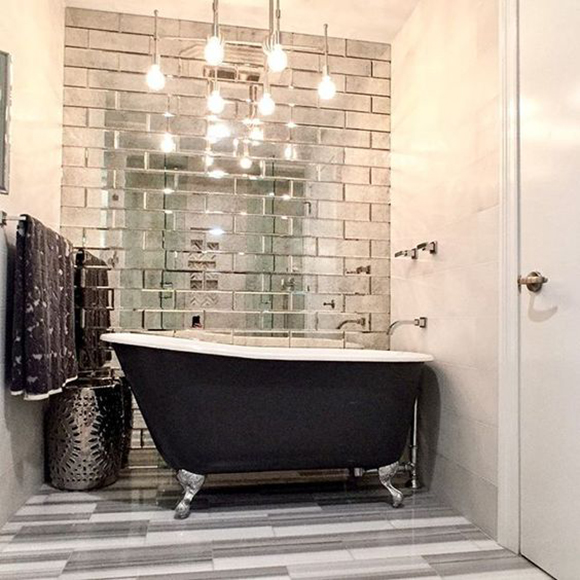 Faux brick mirrored tiles behind a freestanding silver-leaf clawfoot tub painted black in the outside, with silver taps from the wall. Above, multiple round lightbulbs are exposed in a modern chandelier light fixture. On the left wall hangs a black towel on a silver towel rail, and a silver waste bin sits in the corner, atop white and grey horizontal tiled floor. The white door is just visible at the right of the image.