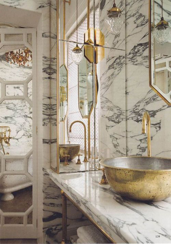 A white and grey marbled  bathroom counter and walls are counterpointed with brassy and silver patina vessel sink with a curved gooseneck faucet. Brass detailing around the counter, mirror, and storage elements match the vessel sink and are reflected in floor-to-ceiling mirrors.