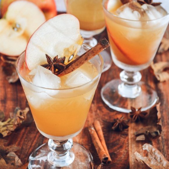 Photo of a cocktail glass with an orange mixed drink, a cinnamon stick, and a clove with a slice of apple and some ice, set upon a dark wood table, surrounded by other glasses in the background.