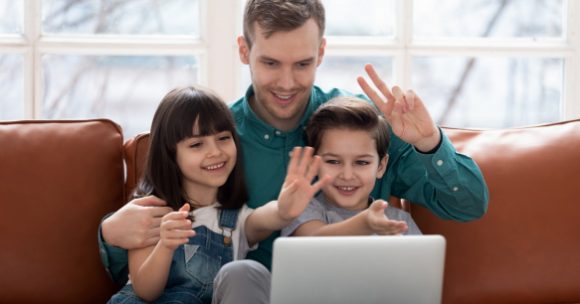 Photo of a father and two young girls waving at a computer screen, sitting on a leather couch.