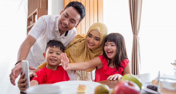 Photo of a multi-cultural family at the dinner table posing together for a cell phone photo: Asian dad in a white shirt, Middle-eastern mom waring a yellow hajib, a boy and a girl both in red shirts.
