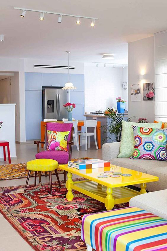 Photo of a light colored open-plan living and dining area, with a bright pink chair, a bright yellow coffee table, a bright yellow footstool, bright red floral patterned rug, a second footstool in multicolored stripes, and multiple pillows in colorful patterns.