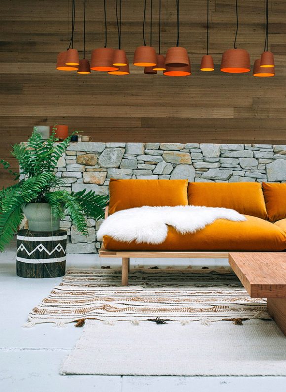 A modern living room with wooden wall at the back, a half height rock wall in front of that, an orange velvet sofa with a white faux fur on top of a patterned rug and a light colored coffee table. To the left of the sofa sits a green plant. Above the sofa hangs a dozen orange wood round pendant lamps.