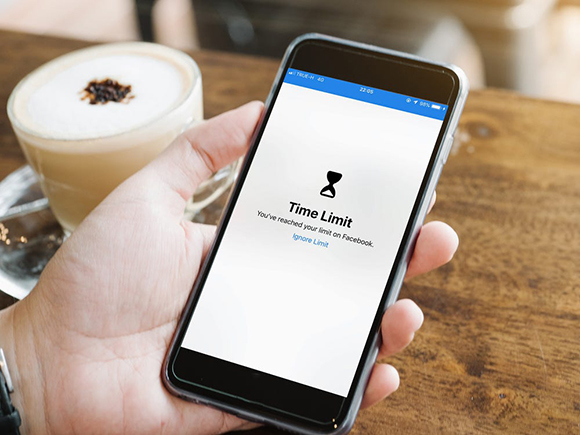 A cell phone in a person's hand with a white screen and timer graphic showing the words "Time Limit - you've reached your limit on Facebook". To the side is a coffee drink.