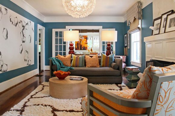 Photo of an eclectic living room, with white ceiling and trim, light turquoise walls, wood flooring with a white shag rug with brown giraffe-lines, a silver and orange patterned chair in the foreground, a light-colored round wood coffee table in the center of the room, a brown 3-seater sofa with multiple mismatched throw pillows in orange and turquoise, plus a table with two matching lamps behind the sofa.
