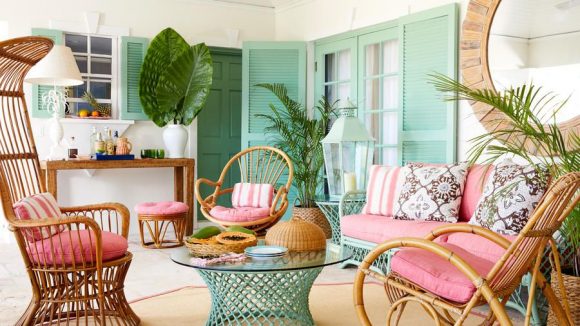 View of a Bahamian living room featuring cane chairs and sofa in pinks, a cane glass-topped table in minty green, and matching green shuttered blinds on the windows. Multiple green plant fronds are scattered around the space.