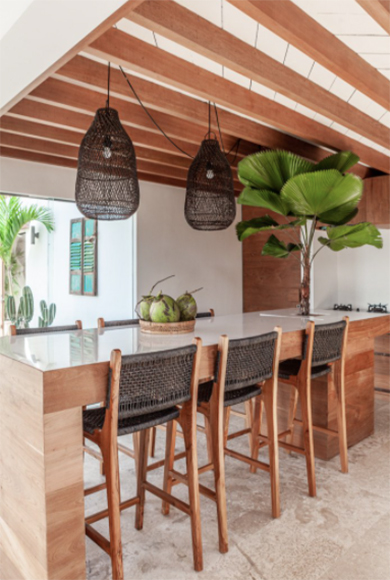Photo of a bright, light-colored kitchen with 3 counter-height chairs in dark rattan with a pair of matching dark rattan pendant lights hung above the white counter. A large palm tree grows from the center of the counter's end cap.