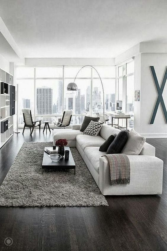A living room, with dark wood flooring, a grey rug in front of an L-shaped grey sofa with black pillows, grey blanket, black coffee table, and in the background, black and white square shelving, near a cityscape window.