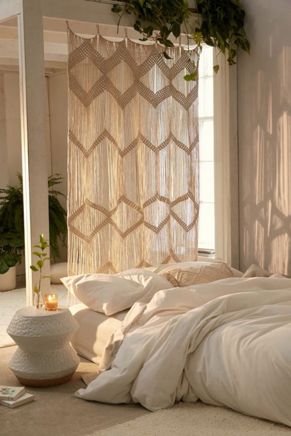A room all in creamy whites, with a rug, under a bed on the floor, with sheets and blankets spilling over. Behind the bed hand a macrame panel with criss-crossed square patterning. Light streams through the panel leaving shadows on the wall.