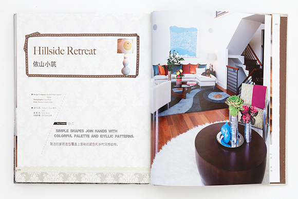 Photo of a 2-page spread of a book, showing the project title Hillside Retreat in English and Chinese, and a colorful living room with round wooden table, fluffy white rug, chair with bright pink throw blanket casually tossed over the back, and in the background, a seating area featuring a sofa with multiple bright colored throw pillows and coffee table on a patterned rug.