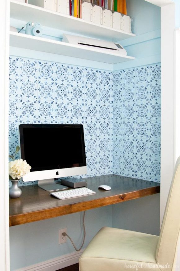 A pantry is converted to a simple pocket office by wallpapering with light blue patterns and matching blue paint on the upper walls. A reddish wooden desk surface is inserted, with a Mac computer and small vase of flowers, in front of a tan chair. Higher up are white shelves with white matching magazine storage boxes, and a small white printer and light blue camera.
