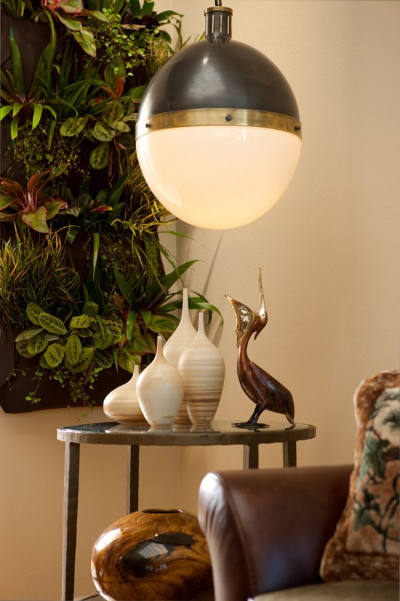 Closeup view of a living room corner, with a peek of a brown leather sofa next to a brown metal round side table filled with multiple vases of varying sizes and shapes, all in white with slight brown stripes. In the corner, a pocket wall of live plants. Above the table, a round white pendant light.
