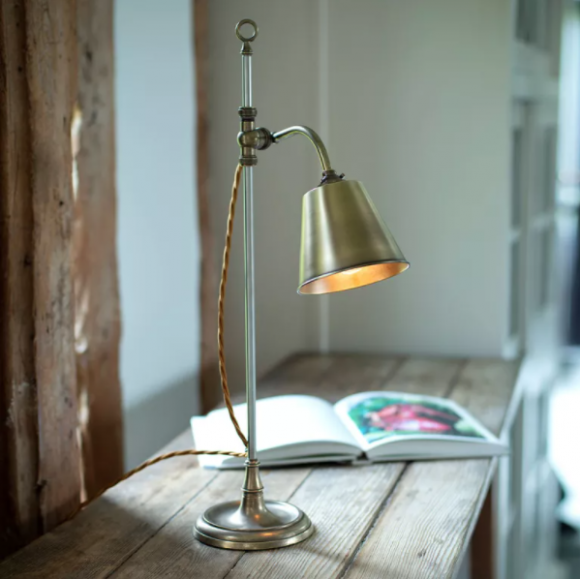 Photo of a simple brass arched desk lamp with an round lampshade and a twisted exposed cord, on a rough wooden table with an open book nearby.
