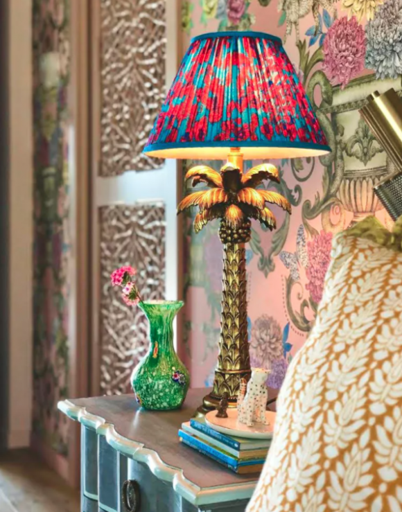 A riot of colors and textures in a hallway: floral wallpaper, carved room divider, a side table stacked with small books, and a lamp made to look like a coconut tree, with a bright blue and hot pink spotted lampshade, next to a yellow and white patterned pillow on a chair.