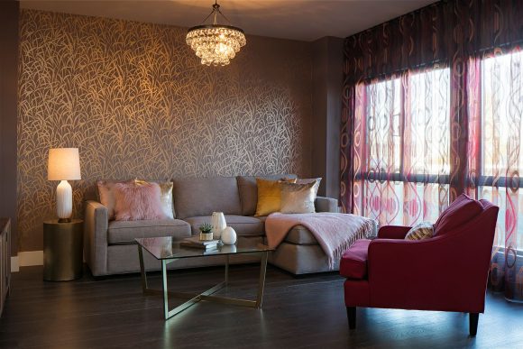 View of a living room with plant-inspired gold on tan wallpaper, a tan sofa with a chaise long, a hot pink armchair, and a glass coffee table, all underneath a four-tiered round glass pendant light that looks like a mini chandelier.