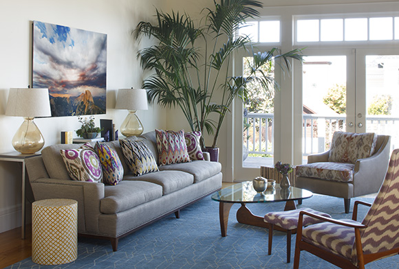 A comfortable and colorful living room, with a blue patterned rug over warm wood flooring, a grey sofa scattered with multi-patterned and colored throw pillows, a large plant in the corner behind. Two chairs and a coffee table create a comfortable conversation corner. A colorful photo is hung on the wall behind the sofa, featuring the sky and mountains.