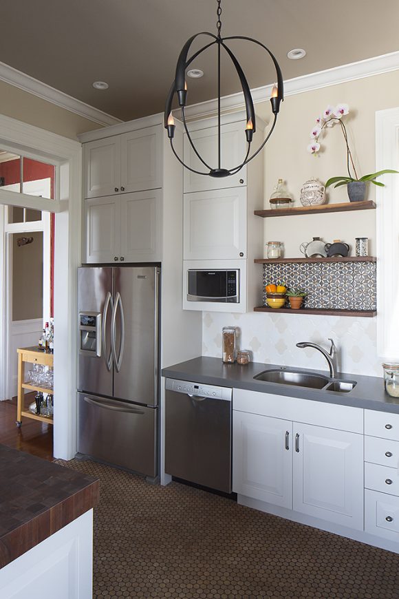 A country-style kitchen with penny tiles, white cabinetry, grey countertops, silver appliances, and tan and white tiled backsplash. Hanging in the center of the space is a pendant light formed from two black metal rings at 90-degree angle, with four black faux candle holders, and a stabilizing single vetical metal pole in the center.