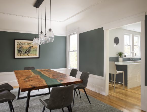 Dark grey-green dining room walls stand out against the white molding and ceilings. 4 glass pendants with exposed bulbs hang above a cypress wood dining table with a green stripe running up the middle, surrounded by 6 modern dining chairs. The white and grey-green kitchen is visible through the doorway, with a peek of the counter seating and windows showing through.