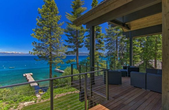 Another view of the dock extending out into the blue waters of Lake Tahoe. Greenery and trees surround a dark wooden deck with black seating, bounded by a modern black railing.