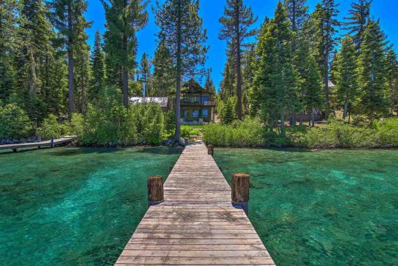 A beautiful view down a long wooden dock towards a home in Lake Tahoe, surrounded by emerald waters and green trees.
