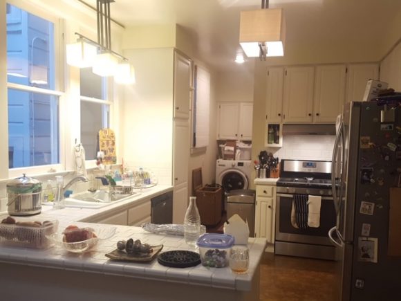 A cramped, dark kitchen without storage space, as evidenced by multiple items stored on the countertops. The cooktop has only a narrow strip of counter next to it. The laundry room is visible at the end.