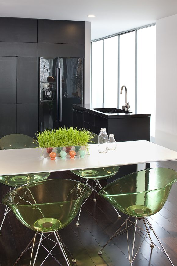 Image of a dark wood bachelor's kitchen with dark surfaces and a gooseneck faucet. In the foreground, a white table is surrounded by green lucite chairs with silver metal legs. On the table as a centerpiece is a planter filled with growing green grass.