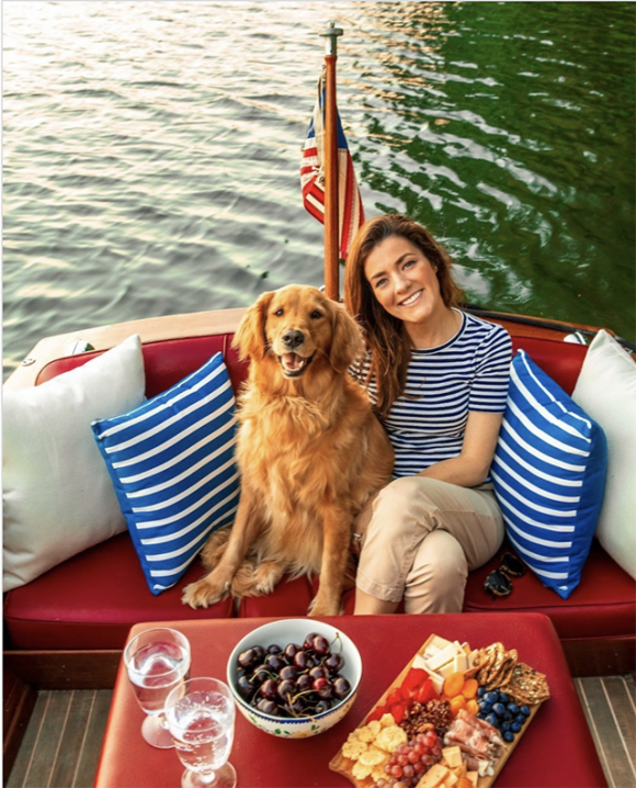 A golden retriever sits next to her owner, who is wearing a blue striped shirt matching the pair of pillows either side of them, on a boat on the water. In front of them sits a cheese and fruit plate and pair of glasses, plus a bowl of cherries.
