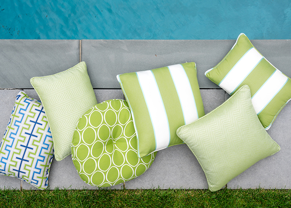 Multiple throw pillows in lime green and white stripes, lime green and white circles, and turquoise blue, white and lime green square patterns are strewn about next to a swimming pool, alongside a swatch of green grass.