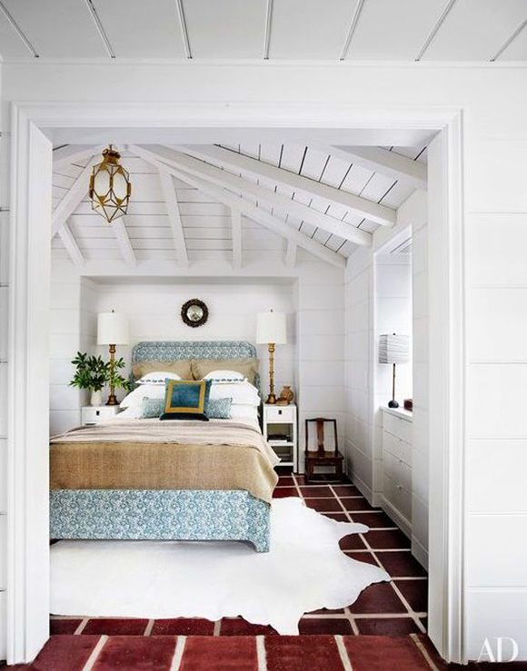 An all-white wooden bedroom with large dark red brick colored floor tiles with white grout covered in a white faux skin rug. A queen sized bed in natural beige colors and blue patterns is topped by a blue and gold square pillow. White table lamps on white side tables flank the bed.