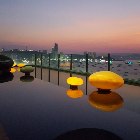 A rooftop view over a Thai city skyline, with clear glass walls between black posts allowing the view in through the protective railing. On the roof, an infinity pool reflects large and small rocks delicately balanced above the water. The stones light up, their orange color matching the sunset beyond.