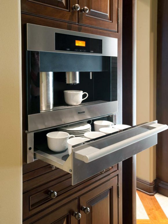 A custom built-in Miehle coffee machine above a partially-open stainless steel drawer filled with white cups and saucers. Dark wood cabinetry is visible above and below.