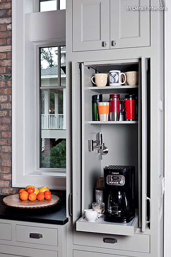 A custom slide-out with a small coffee machine, creamer and sugar, underneath a shelf of colorful mugs, next to a window with a view to the outside. White cabinetry with black countertops, silver and dark metal drawer pulls and handles are visible next to a small stripe of exposed brick off to the left of the window. A plate of oranges dresses the counter.