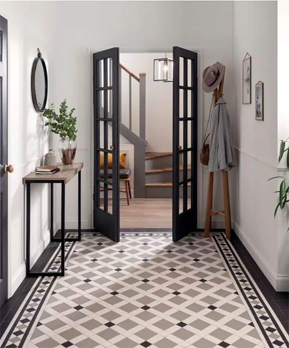 An entryway featuring latticework criss-cross tiling in tan, black, and white, leading to partially open black french doors into another hallway with wooden floors. In the main entry you see a console table dressed with plants and books, a free-standing hat and coatrack, a hint of green plant off the right, and mirrors and artwork hung on walls either side.
