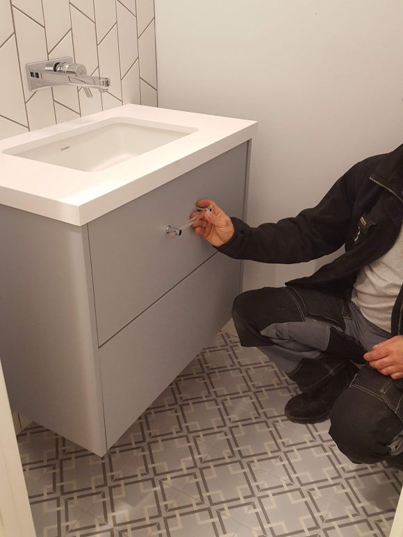 Our contractor kneels and holds up a glass drawer pull to the front of the grey cabinet under a white basin sink. The ceramic tile flooring is a series of square geometric patterns in white and dark grey on a light grey ground.