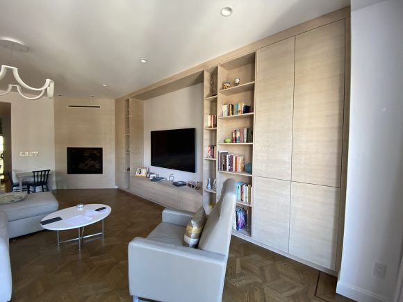 A living room with medium brown parquet wood flooring, light wood custom cabinetry with open bookcase shelving, a wall-mounted television, a modern custom fireplace on the far wall, and grey armchair and corner sofa with an oval coffee table between them. Recessed lighting peeks through a white painted ceiling, and a modern geometric swoop chandelier hangs above the seating area.