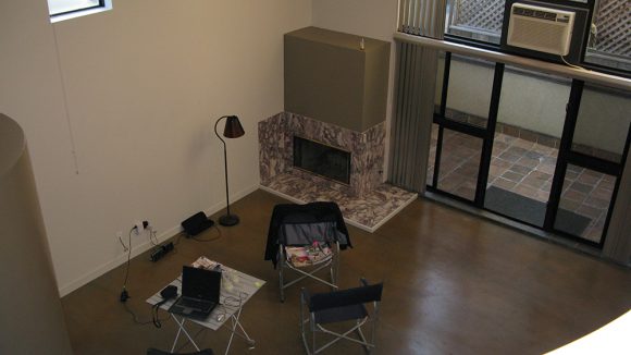 A dated-looking moss-green boxed fireplace has a multi-colored surround and black fireplace insert, flanked by black folding chairs and a white folding table, a black standing lamp, and an old window air conditioner above the door to the patio.