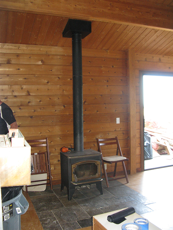 A tiny black potbelly stove sits on slate flooring in front of knotty wood walls with a cabin feel. Two folding chairs flank the stove.