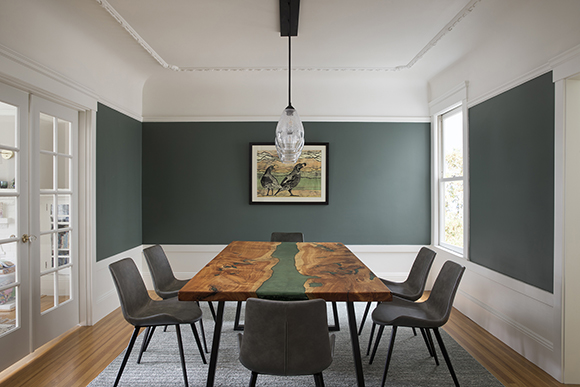 A grey-green wall paint counterpoints white ceiling, chair rail, windows and doors. Grey dining chairs surround a cedar wood table with a stripe of matching grey-green up the middle. A row of clear glass pendant lights hang above the table, and at the back wall in the center, a picture of two quail in colors that tone with the grey green and warm wood.