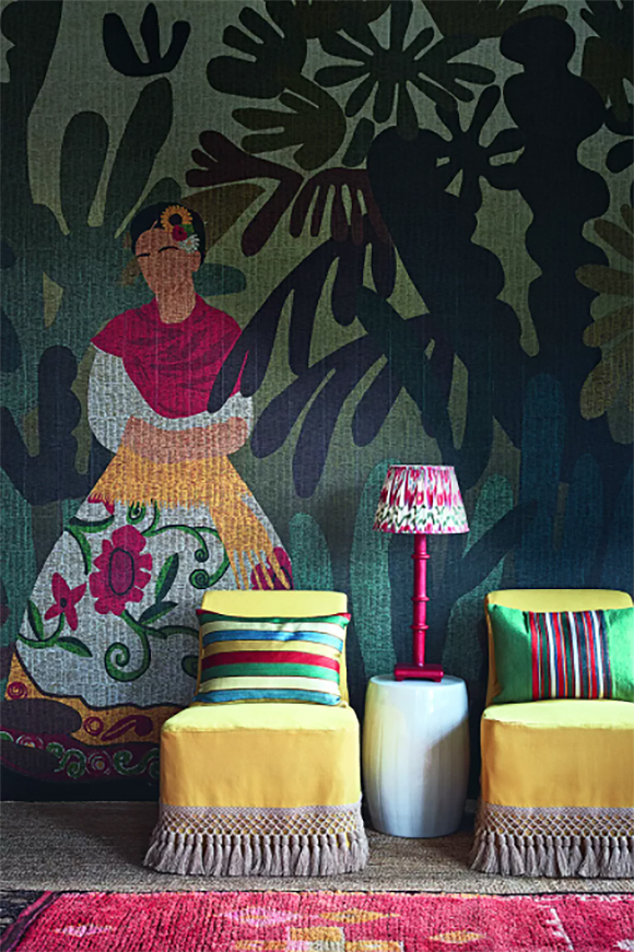 A green and blue fabric wall mural features a white, yellow, pink, and green dress on a Spanish woman wearing a pink shawl and 3 flowers tucked behind her ear. In front of the mural are two yellow chairs with hand crotched tassels at the bottom, and two multicolor striped pillows on the chairs. Between the chairs sits a small white table with a pink lamp.