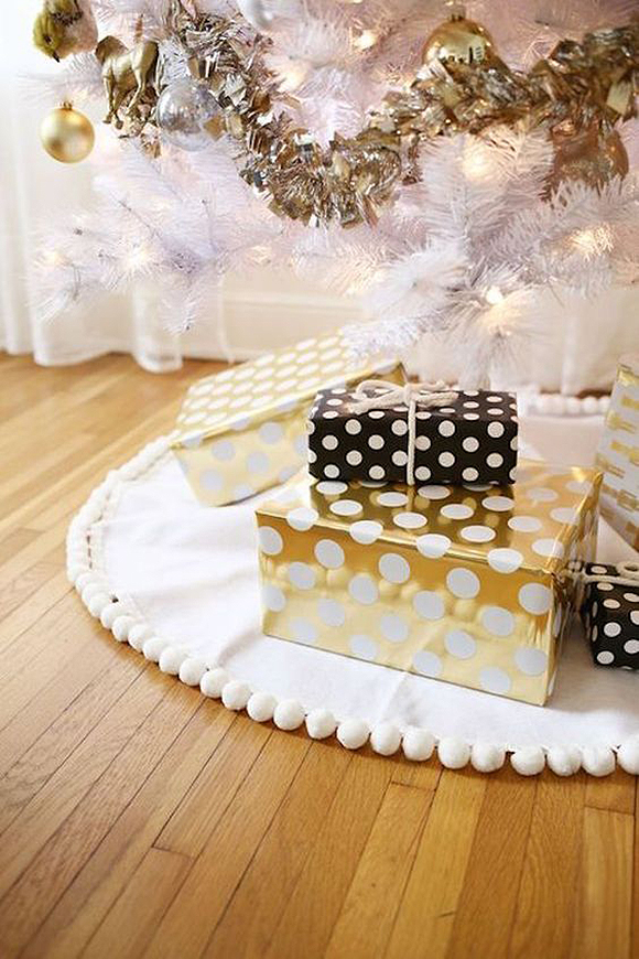 A stack of gifts under a white Christmas tree with gold garlands, on a white surround under the tree, wrapped in black-and-white polka dot and gold-and-white polka dot paper.