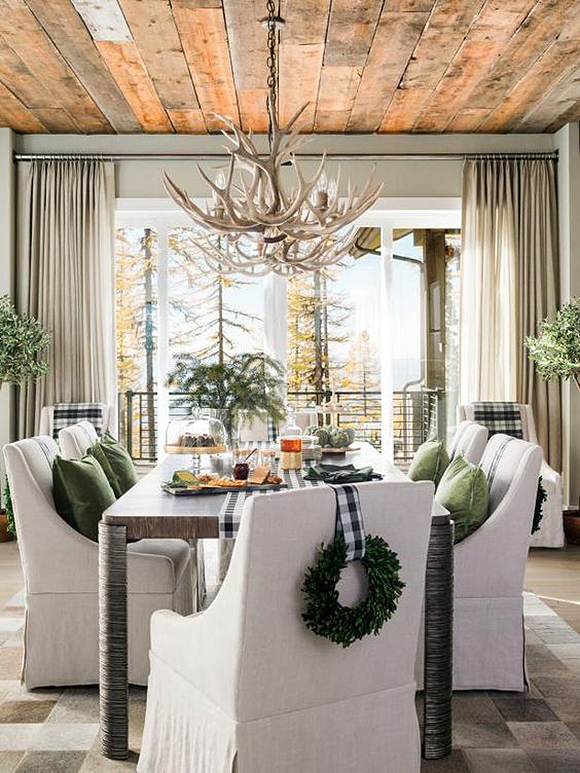 Holiday tablet wet with black and white checked table runner, cheese, dessert and wine. The 6 white dining chairs are dressed with a green pillow on the seat and a green wreath hung over the back with black and white checked fabric that matches the table runner. Green plants are scattered around the room. Overhead, a deer antler chandelier continues the outdoor touches.