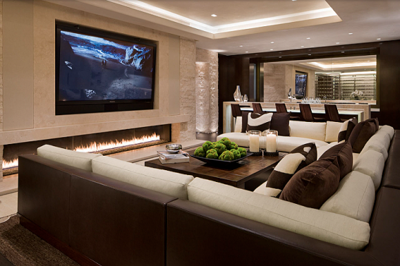 A brown and white corner sofa under a sunken ceiling with recessed lighting, in front of a large TV screen, and a ribbon fireplace underneath. Across the room is a wine bar with counter-height seats matching the sofa colors.