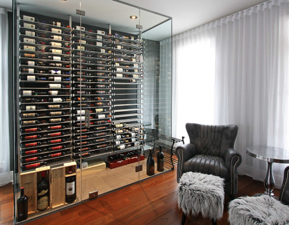 Enclosed glass room-within-a-room featuring dozens of wine racks stored on their side, label out. Outside the glass room, a leather chair and fuzzy ottoman sits next to a glass pedestal table, for resting your wine glass.