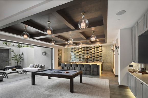 An open-plan room with a billiard table in the center, a wine bar with counter-height seating, a living room area to the left, and on the right, a kitchen space.